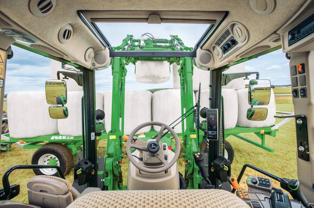 A John Deere traktor fülkéjét látjuk a vezető szemeszögéből, amint épp csomagolt bálákat pakol a homlokrakodóval
